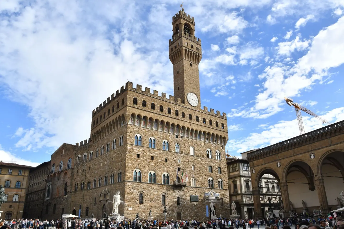 Gran palacio medieval de piedra con almenas y una alta torre con reloj, en una plaza llena de gente con estatuas blancas al pie del edificio, bajo un cielo azul con nubes y una grúa a la derecha