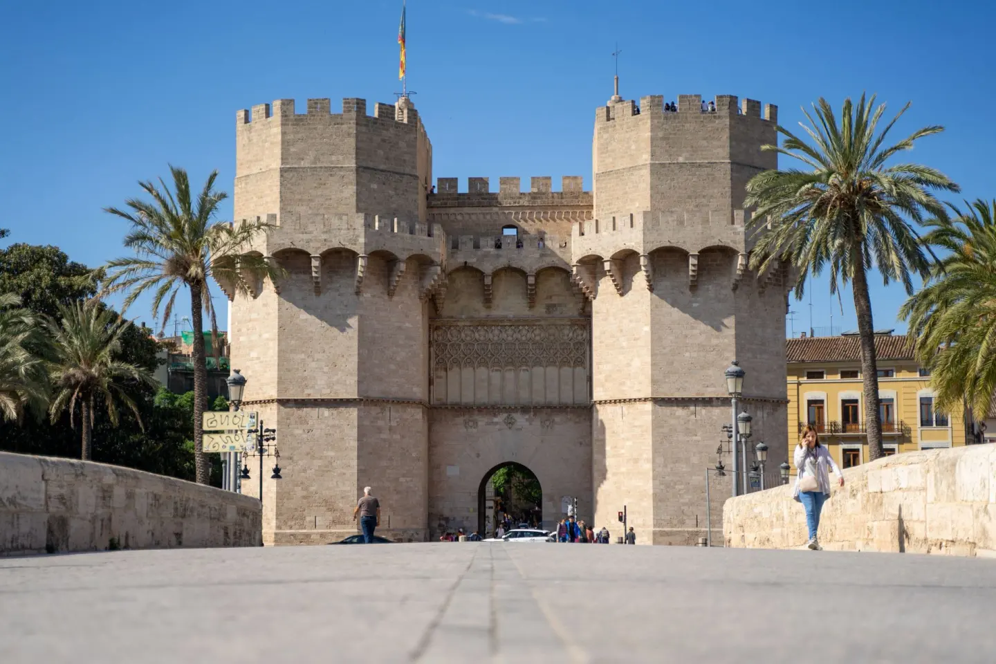 Puerta fortificada de piedra con dos torres almenadas y un arco central, vista desde un puente con palmeras y gente caminando