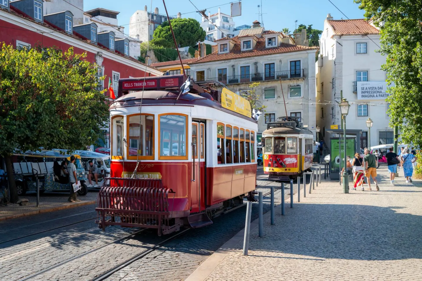 Dos tranvías antiguos, uno rojo y blanco y otro amarillo, circulando por una calle empedrada entre edificios de colores, con gente caminando por la acera