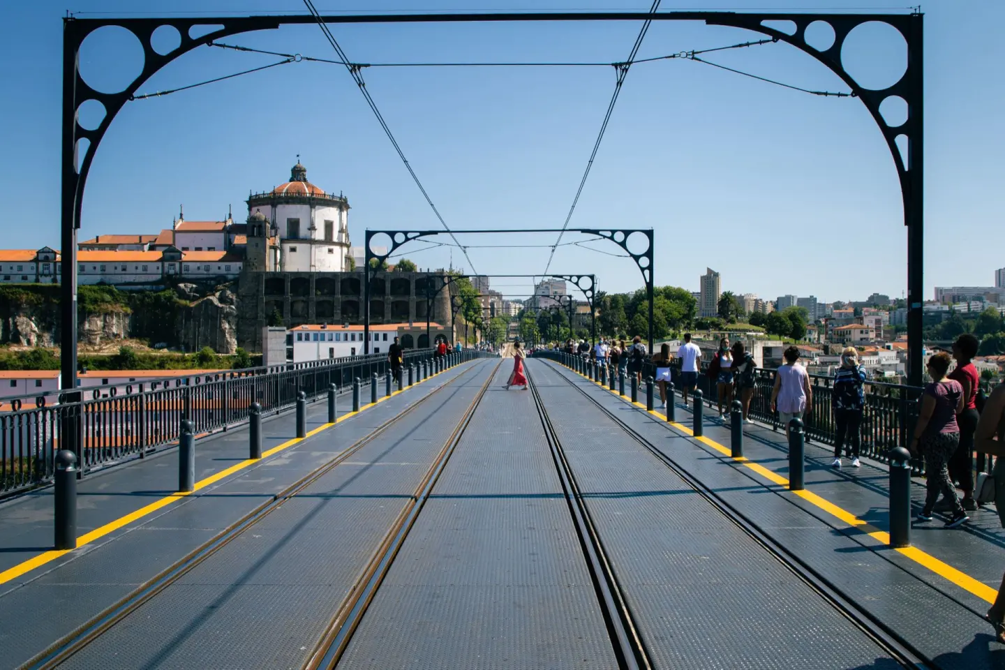 Tablero de un puente metálico visto de frente, con vías de tren que se pierden en la distancia entre arcos de hierro negro, gente caminando por los lados y, al fondo, un edificio blanco con cúpula sobre una colina, bajo un cielo azul