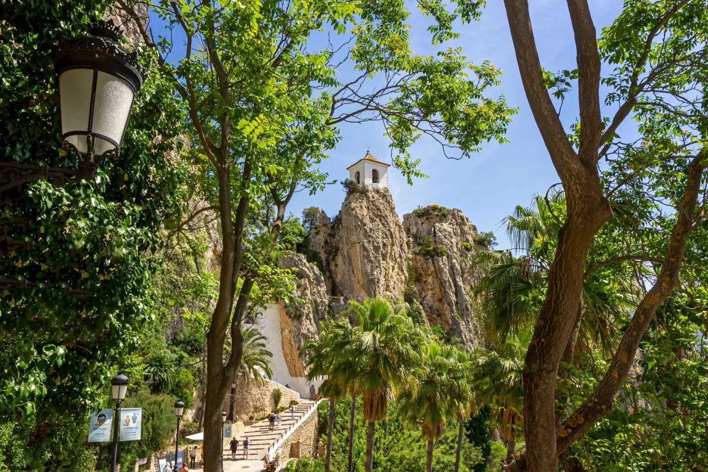 Campanario blanco sobre un peñón visto entre árboles y palmeras, con una escalera de piedra que sube por la ladera