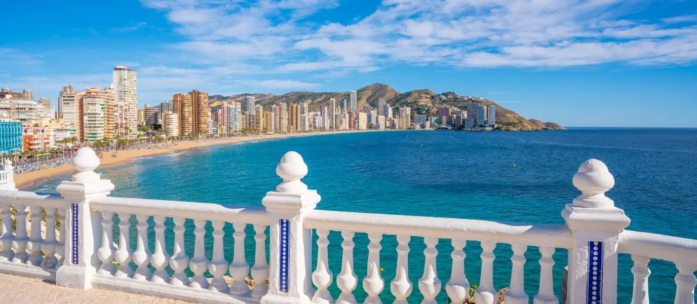 Balaustrada blanca en primer plano y, al fondo, una playa de arena con rascacielos junto a un mar azul turquesa