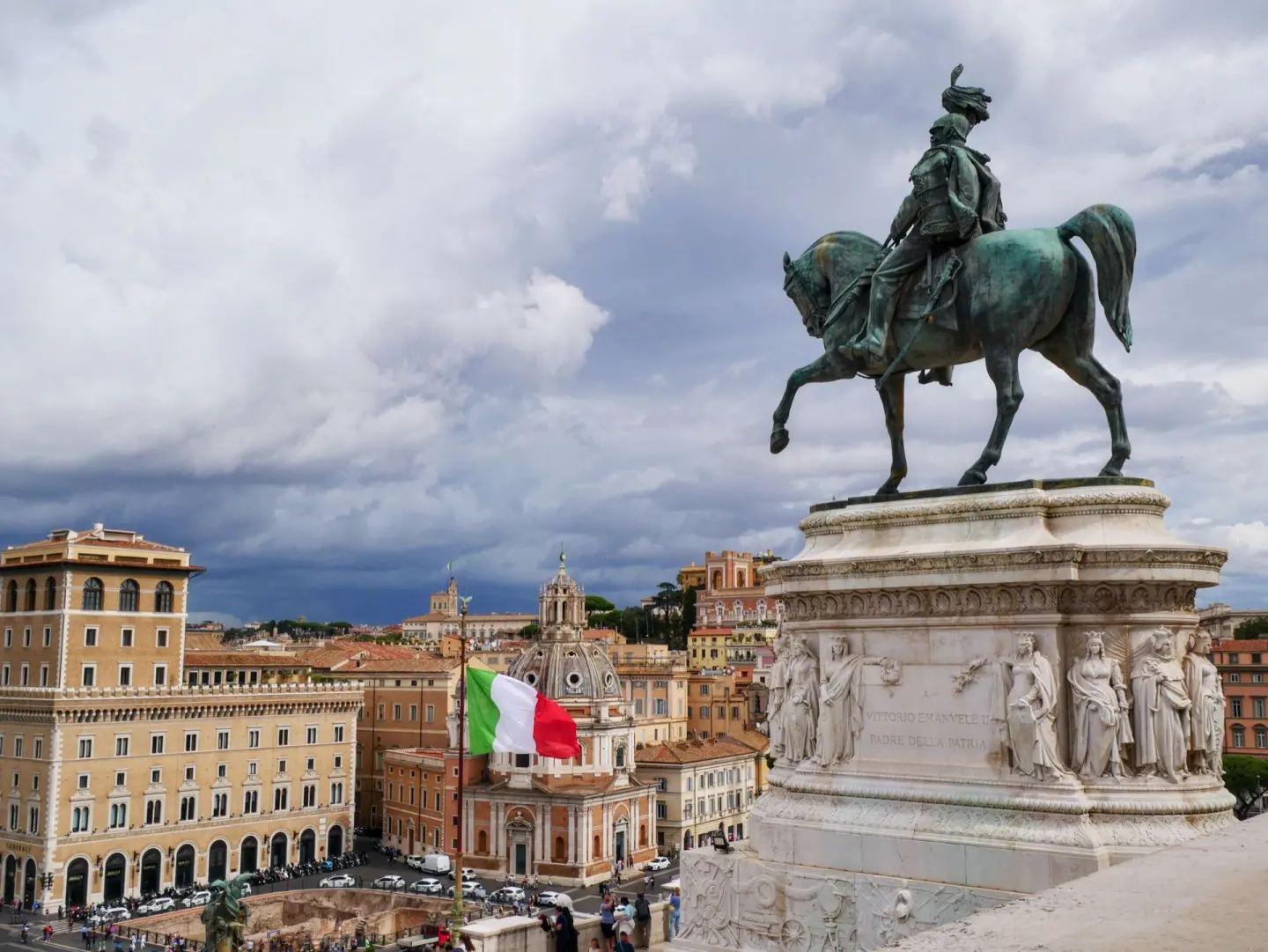 Estatua ecuestre de bronce verdoso sobre un gran pedestal de mármol con relieves, con una bandera de Italia y una plaza con edificios de tonos cálidos bajo un cielo de nubes oscuras